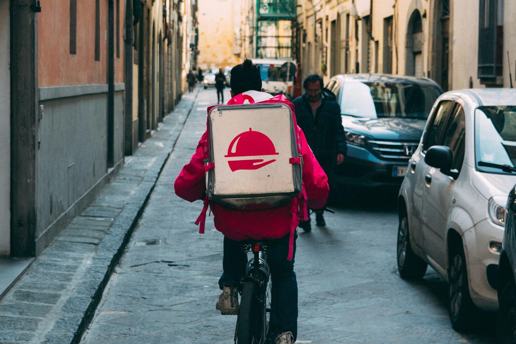 A food-delivery courier riding a bicycle down a narrow city street with a delivery backpack
