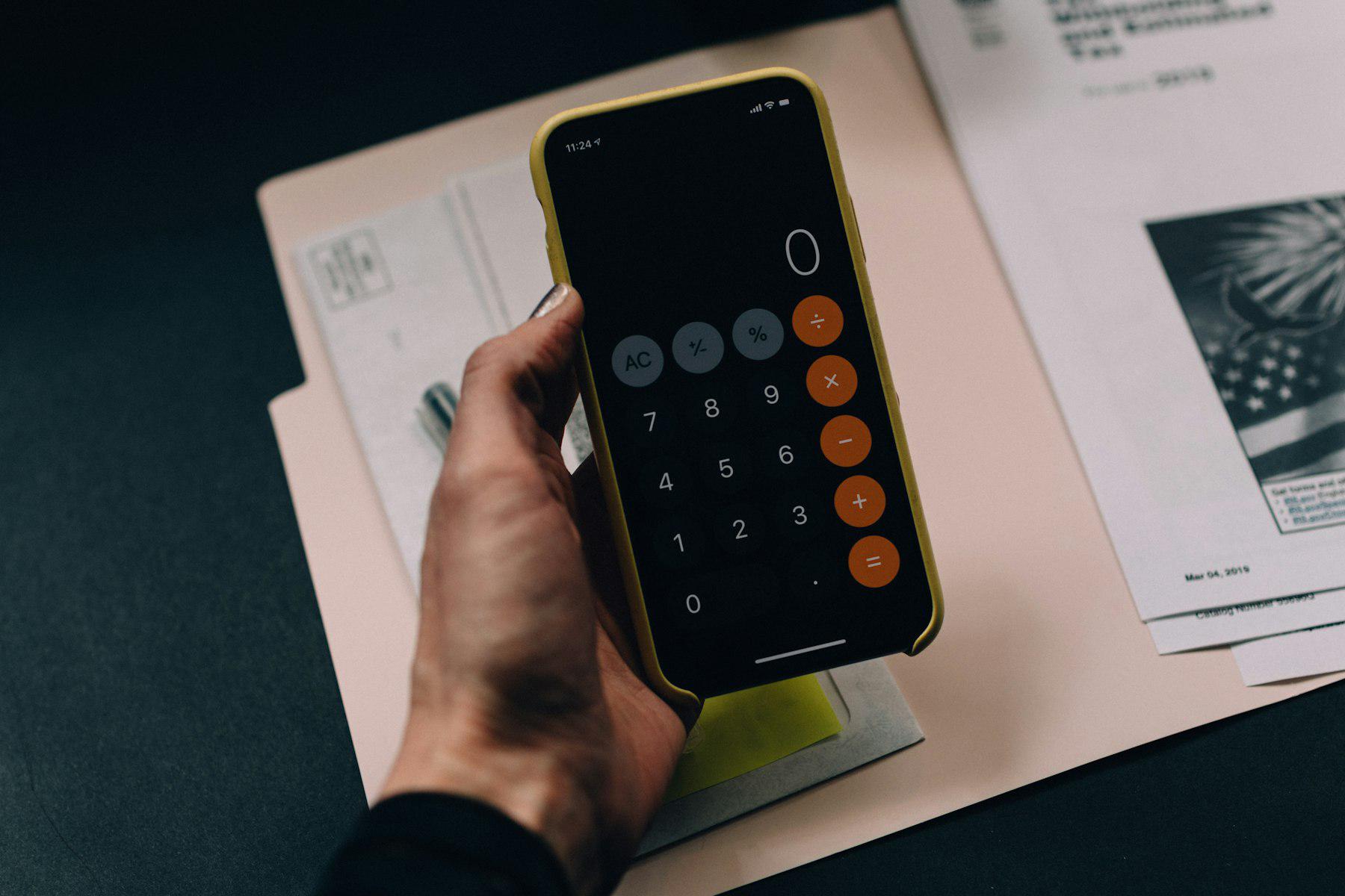 A hand holding a smartphone calculator app over tax documents on a desk